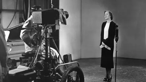 A black and white image from 1936 - a smartly dressed lady stands in front a microphone and a television camera 