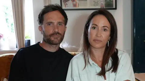 Qays Najm/BBC Craig and Carly Bowe sitting on a sofa. Craig is wearing a black T-shirt. Carly is wearing a turquoise shirt and has long brown hair.