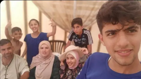 al-Dalou family photograph A selfie taken by Sha'aban al-Dalou showing him and his family. He is a young man of 19 in a blue t-shirt, standing in the foreground, with six relatives aged from childhood to middle age smiling behind him. Some of the children are making thumbs-up gestures.