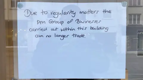 A white sheet of paper stuck to glass. On it there is black writing which reads: "Due to regulatory matters the PM Group of Businesses carried out within this building can no longer trade."