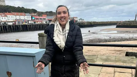 A woman wearing a black coat and pale scarf holds out her outstretched muddy hands. Behind her is the beach in Whitby.