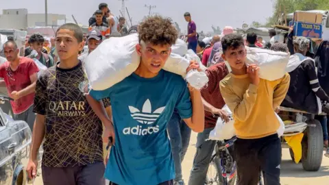 Three young men are pictured with food supplies in sacks on their backs, as part of a crowd of people collecting food in the background