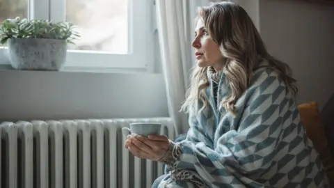 Getty Images A woman wrapped in a shawl sits next to a radiator