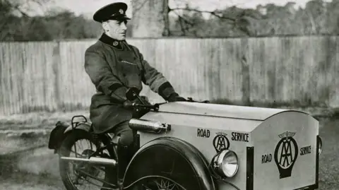 The AA A black-and-white photo of a three-wheeled motorcycle bear the AA logo and being driven by a man in uniform.