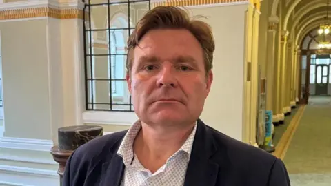 A man wearing a white shirt with small blue pattern and suit jacket. He is standing in a corridor that is on the top of a landing and has green carpet as well as a door at the end. He has short brown hair with a side parting.