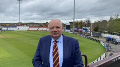 Northamptonshire County Cricket Club Gary Hoffman wearing a blue suit, blue shirt and claret and yellow striped tie. He is stood in the stands of Northamptonshire cricket ground with the green pitch in the background.