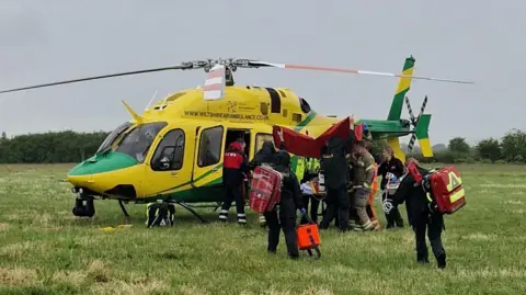 David Bernstein A Wiltshire Air Ambulance has landed in a field. Paramedics and fire crews are seen gathered around the helicopter.