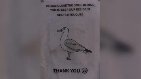 Stuart Harmer a paper poster with a picture of a seagull and the message "please close the door behind you to keep our resident shoplifter out! Thank you.""