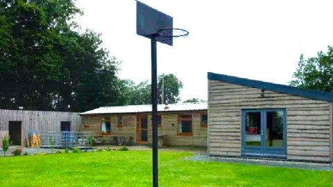 Cheshire East Council One of the homes built without permission. In the foreground, there is a lawn that has a basketball hoop on it. The single-storey wooden building, which is in two sections has a slanted roof.