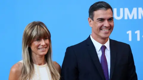 Reuters Sánchez and his wife Begoña Gómez are smiling with a blue background. Sánchez is wearing a suit and Gómez a white dress.