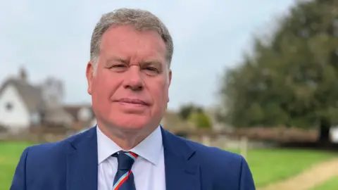 Sam Read/BBC Greg Horsford standing outside looking directly at the camera. He is wearing a navy blue suit jacket, white shirt and blue and red patterned tie.