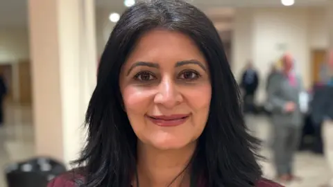 Shariqua Ahmed/BBC Shabina Qayyum with black shoulder-length hair wearing a burgundy jacket, smiling for camera.