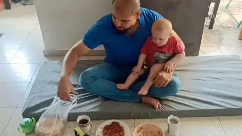 Dr Elawa sits cross-legged on a floor mat on a tiled floor. A small child in a red t shirt and with fair hair sits on his knee. He is leaning towards two dishes of food in front of them. There is also a toddler's drinking cup.