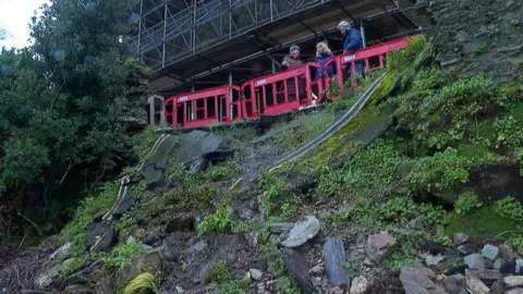 A landslip on a road in Dartmouth. There are red plastic fence barriers at the top of the landslip, where there are three people stood talking.