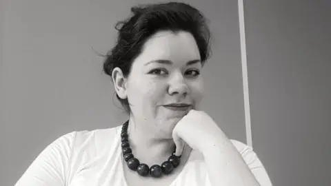 Rachael Clarke Rachael, who has dark hair, wearing a white t-shirt and a chunky bead necklace looks into the camera while resting her chin on her hand. The image is a black and white photo.