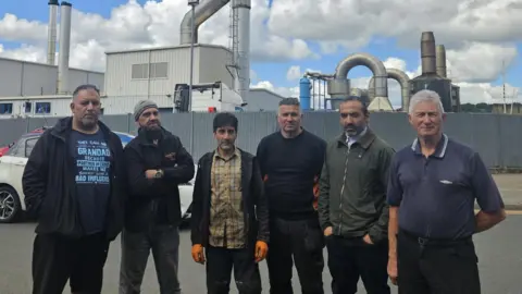  A group of 6 men, of varying ages and background, stand in front of a factory plant. They are on a road, with cars parked around them and passing behind them. They look quite serious. Some are dressed in workman like clothes.
