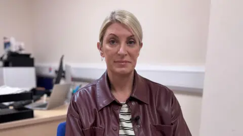 Linda Dillon. She has blonde hair tied back, wearing a brown leather shacket and black and white striped top.