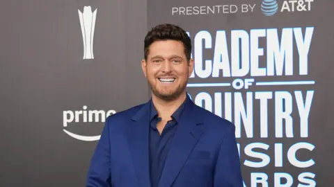 Reuters A man, Micheal Bublé, he is wearing a navy suit, with a navy shirt underneath. He is at a premiere, behind him is a media wall with an advertisement for Prime. 