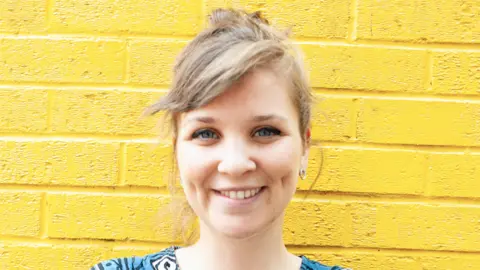 Fumble A woman with tied-up blonde hair stands in front of a yellow brick wall smiling at the camera. 