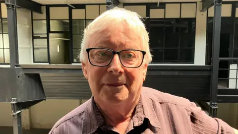 Steve Jones/BBC An elderly man white white hair and black rimmed glasses. He wears a neutral expression while looking at the camera. Behind him are empty rooms used for filming.