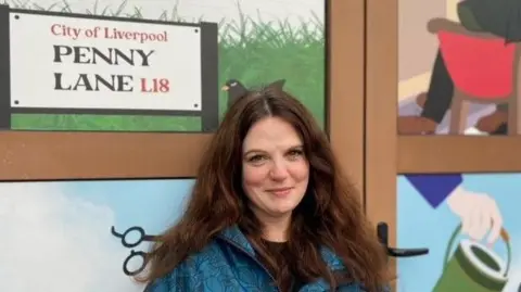 Susannah-Rachel Gorman, wearing a blue zip-up jacket and with long brown hair, smiles as she poses for a photograph in front of her Beatles-inspired Penny Lane artwork.