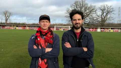 Jonathan Kelham, left, is a white man with brown hair. He is wearing a black cap, a blue raincoat and a black and red Brislington FC scarf. Ken Borg is wearing a blue coat and a black jumper. He has black hair and a short beard. They are both crossing their arms and facing the camera. They are standing on the grass football pitch with a colourful border and a white goal behind them in the distance.