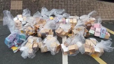 Several plastic bags fill of boxes containing vapes. They are piled upon a floor with white and yellow lines painted on it.