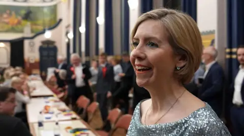 Andrea Jenkyns has short light-brown hair and wears a sparkly blue top. She is smiling as she stands in a large room full of people in formal clothes during an election count.