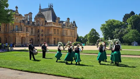 Fiddlesticks A troupe of eight female clog dancers on a lawn in front of a large stately home. They are wearing calf-length green skirts, black waistcoats and white, short-sleeved collared shirts. They are jigging and holding garlands. They have two males drummers with them. It is a sunny day.