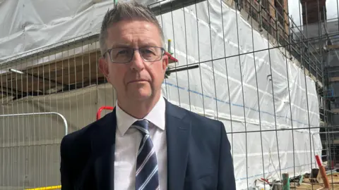 Julia Gregory / BBC A man in a black suit wearing a striped tie. He is stood in front of a construction site with a metal railed fence surrounding it.