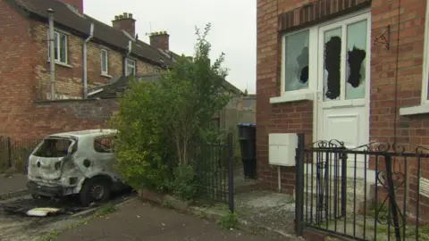 BBC Burned-out car and broken window of house