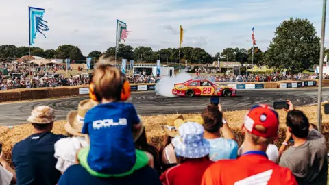 CarFest A crowd watches a racing car on a track at an outdoor festival, with hay bales, flags and spectators in the background.
