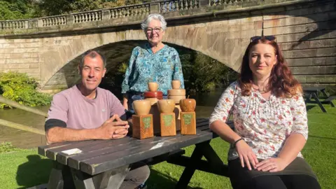 BBC A wooden picnic bench sits on the grass beside a stone bridge. Sat at either side of the bench is a man in a lilac t-shirt and a woman in a flowery blouse. Stood at the end of the table is woman in a flowery green blouse, with short grey hair and spectacles. There are five wooden trophies between 25-35cm carved into an apple on a plinth.