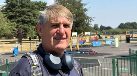 Kevin Shoesmith/BBC A man in his 60s with greying brown hair has large earphones around his neck and a backpack on. He is wearing a purple t shirt.  