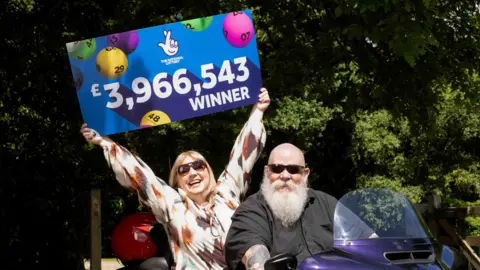 A woman holding a sign that says "The National Lottery and £3,966,543 winner". The background of the sign in clue and there multicoloured bingo balls. There is a man sat in front of the woman on a purple Harley-Davidson trike bike. There are trees behind them and a wooden fence.  