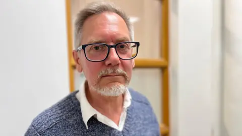 Steve Clay looking at the camera. He has grey hair and a beard, and has glasses. He is wearing a knitted blue and white jumper over a white shirt with collar. Behind him is a white wall with what appears to be a wooden ladder.