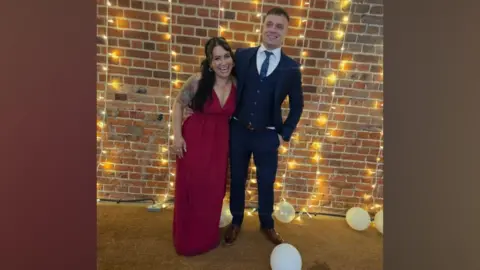 Supplied A woman wearing a red dress stands next to a man wearing a suit with a waistcoat and tie in front of a brick wall