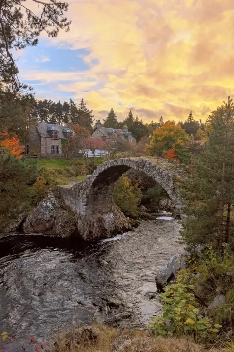 Ellie Berbec The sky is yellow and hangs over a stone arched bridge which has a river running under it.