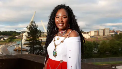 Councillor Lilian Seenoi-Barr stands on the balcony of the Guildhall in Derry City, with the Peace Bridge behind her. The Peace Bridge is white and the sky is cloudy and blue. Lilian is wearing a white off the shoulder long sleeved dress that becomes red and the waist. She has an embroidery necklace with a dozen or so silver charms hanging from it. She has a mayoral necklace as well on that is gold and the emblem is green with a blue river running through it. She has curly long hair and brown eyes. She is wearing lipstick. 