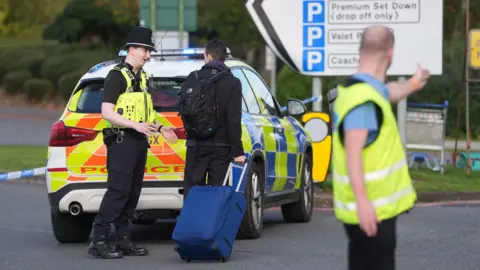 PA media A man pushing a suitcase speaks to a police officer standing in the back of a police car, while another man wearing a high-vis tabard can be seen with his arm outstretched, appearing to give instructions.