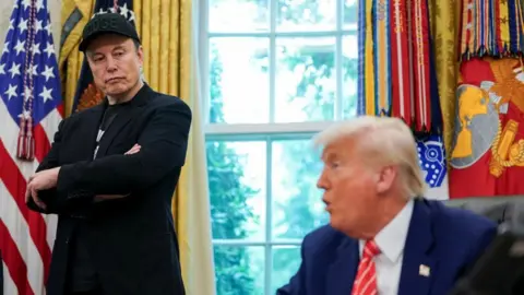 Elon Musk in black outfit stands and looks to his left at Donald Trump who is sitting and talking. An American flag is in the background