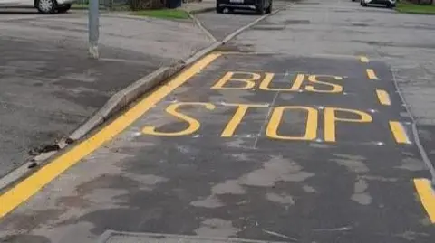 A bus stop with yellow marking on it, with a kerb to the left. Cars are on the road but only their wheels can be seen, in the distance. 