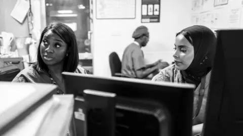 James Clifford Kent Two members of staff, one wearing a hijab, sit at a computer desk in a hospital office environment, looking at a screen while discussing work during a shift change.
