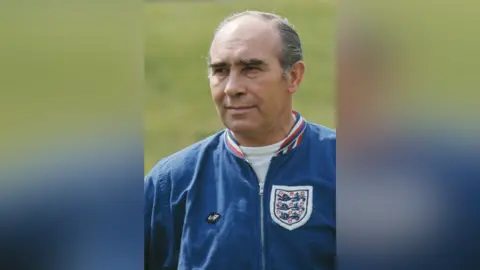 Getty Images A coloured photo of Sir Alf Ramsey on a football pitch. He wears a blue jacket with the England football logo on the chest. He has grey hair which has been slicked back. 