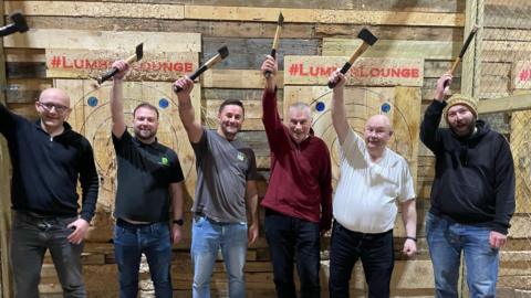 A group of six men at an axe-throwing centre. They are standing in a line and holding their axes in the air, with smiles on their faces.