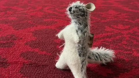 Roo on the carpet at Buckingham Palace - it's a teddy bear on that looks like the baby kangaroo character from Winnie-the-Pooh. It has scruffy grey and white fur with big ears and a long tail. He has a white underside