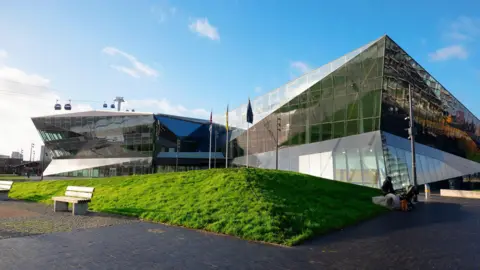 Exterior of the glass covered London City Hall building with grass in front of it and the cable car in the distance