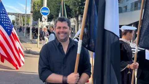Screen Cornwall Leigh Frost is wearing a black button-up shirt, and has black hair and olive toned skin. He is holding a black and white Cornish flag in his hands, and has a brown leather strap on his wrist. A bus stop can be seen in the background of the image, as well as an American flag and many people walking along the pavement.