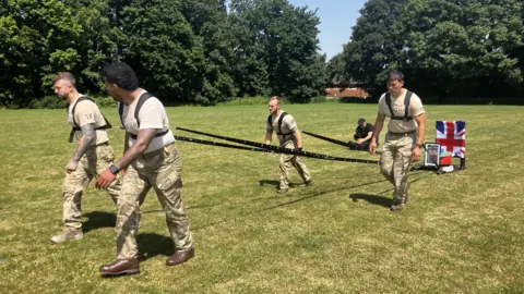 A group of four men wearing camouflage military trousers and beige T-shirts. They are pulling a sled along a grassy field on a sunny day. The sled is decorated with the Union flag and a picture of Andrew Borthwick who took his own life following a battle with PTSD. 