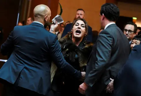 Victoria Jones/REUTERS Two men in suits hold back a  shouting woman with long brown hair. She has her right hand up, holding a folded piece of paper, and is  wearing a traditional possum skin cloak.